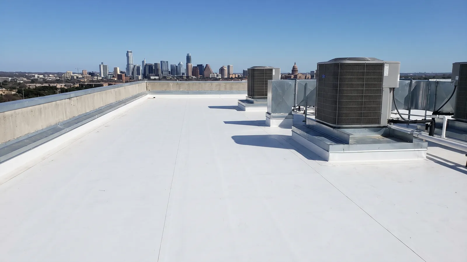 Wide white membrane roof on a commercial building with rooftop air-conditioning units and a parapet, Austin's skyline on the horizon
