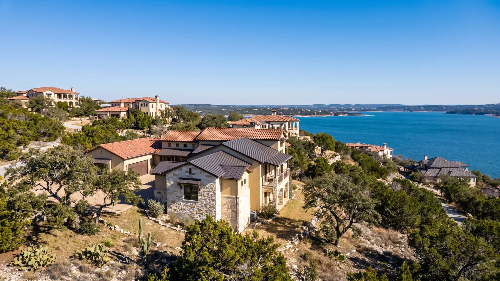 Rooftops of stucco and stone homes on a hillside above Lake Travis near Lakeway, Texas, cedar-covered hills and blue water beyond