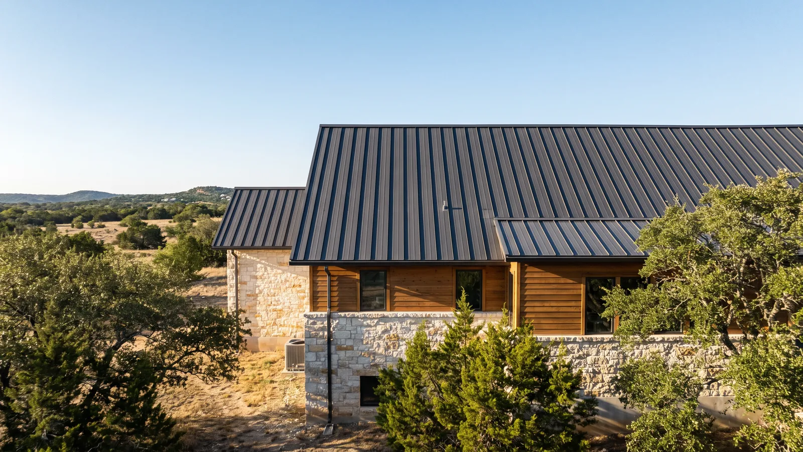 Charcoal standing-seam metal roof on a modern limestone and cedar home in the Texas Hill Country, cedar elms in the yard